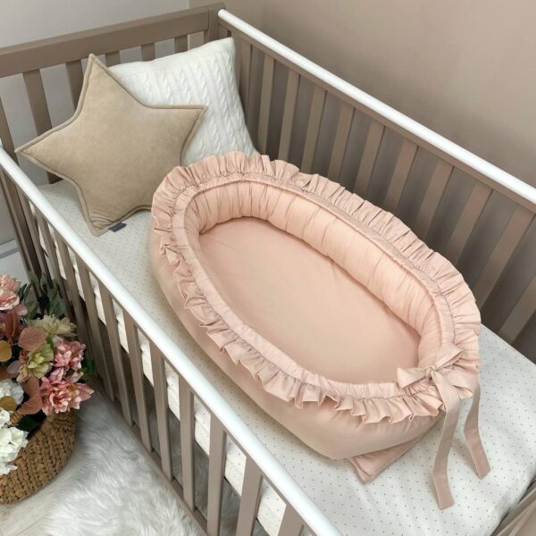 Baby nest cu volănașe în nuanță crem caldă, cu fundă la picioare, fotografiat într-un pătuț