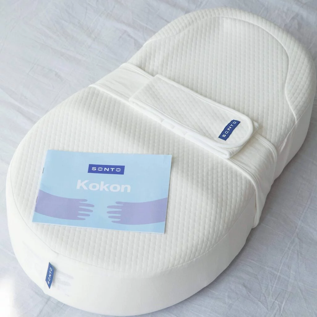 Cocon Sonto Ergonomic Baby Cocoon Cocon Sonto Baby Cocoon alb, matlasat, cu bandă textilă și broșura Kokon, pe un cearșaf