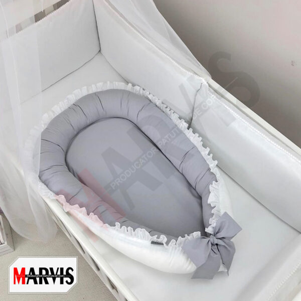 Baby nest gri cu volan și exterior alb, cu fundă gri, așezat într-un pătuț alb