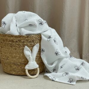Păturica din muselină Mickey Mouse alb revărsată dintr-un coș împletit, lângă un inel din lemn cu urechi de iepuraș