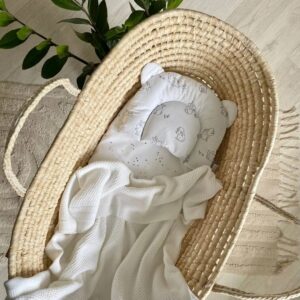 Păturica Cosy albă într-un coș împletit din fibre naturale, lângă o pernuță cu urechiușe