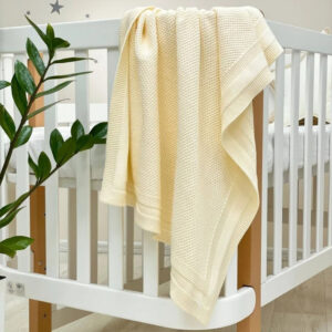 Păturică tricotată Cosy milky, alb lăptos, drapată pe marginea unui pătuț alb cu stâlpi din lemn