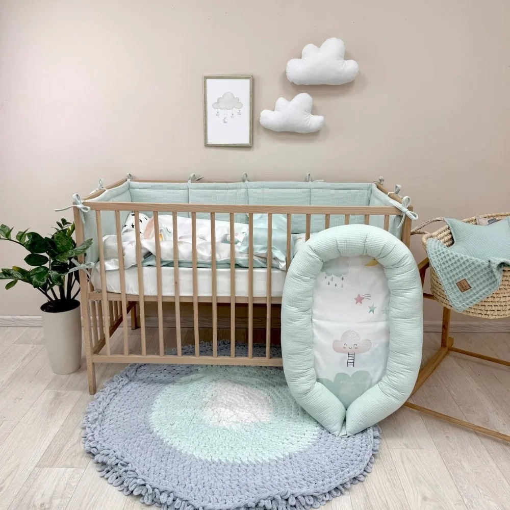 Set lenjerie pătuț Rainbow unicorn Set Rainbow unicorn pe un pătuț din lemn natur, cu apărătoare mentă, lângă un cuib pentru bebeluși