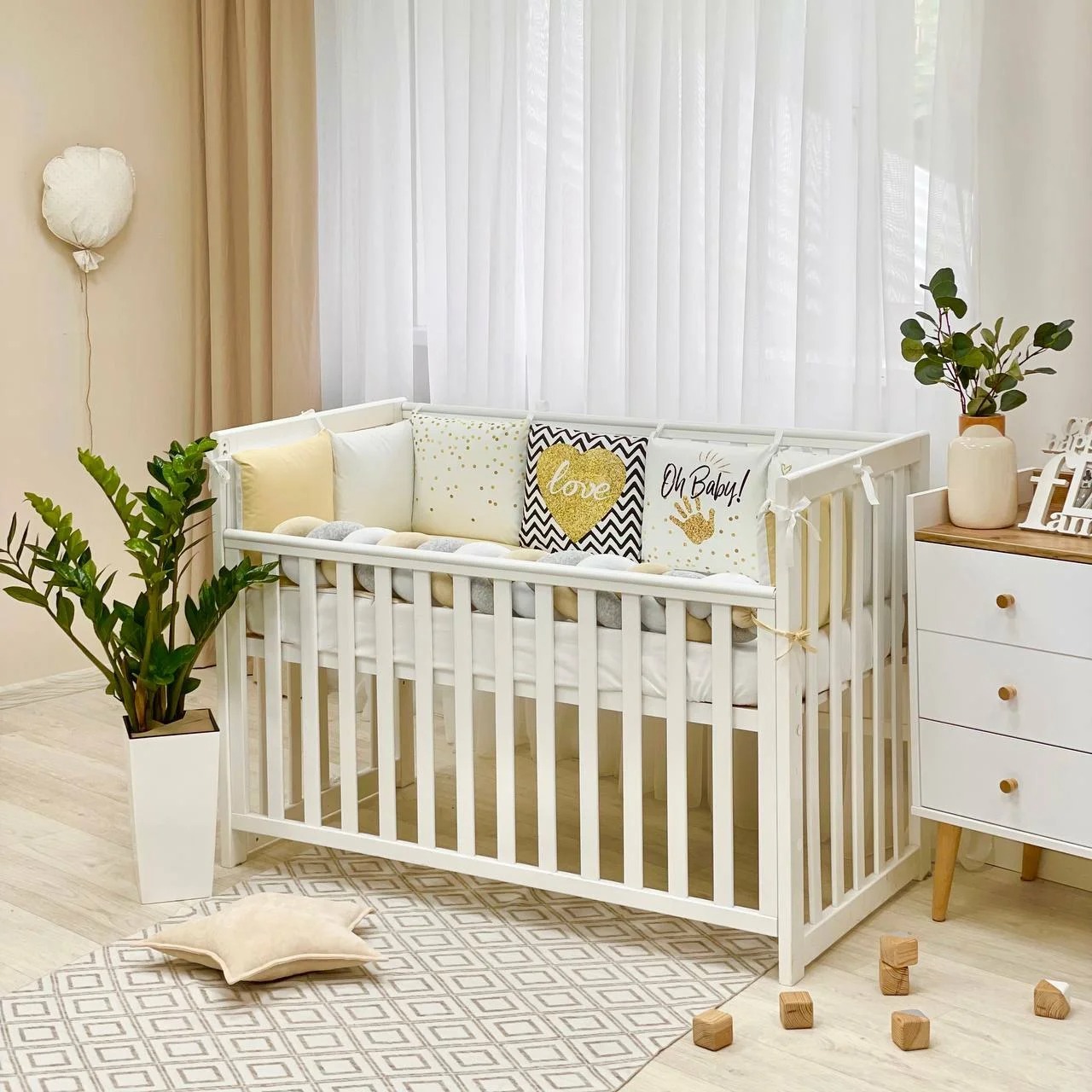 Set lenjerie pătuț Oh Baby, 7 piese Set Oh Baby pe un pătuț alb lângă o comodă, cu pernuțe cu inimă aurie și inscripția Oh Baby