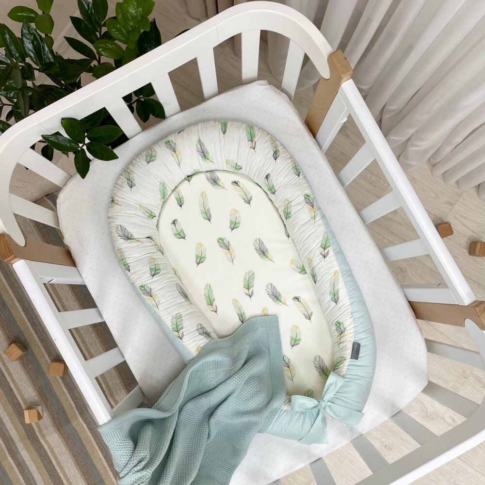 Baby nest Feather mentă Baby nest Feather mentă cu pene verzi, galbene și gri pe fond crem și fundă mentă, în pătuț, vedere de sus