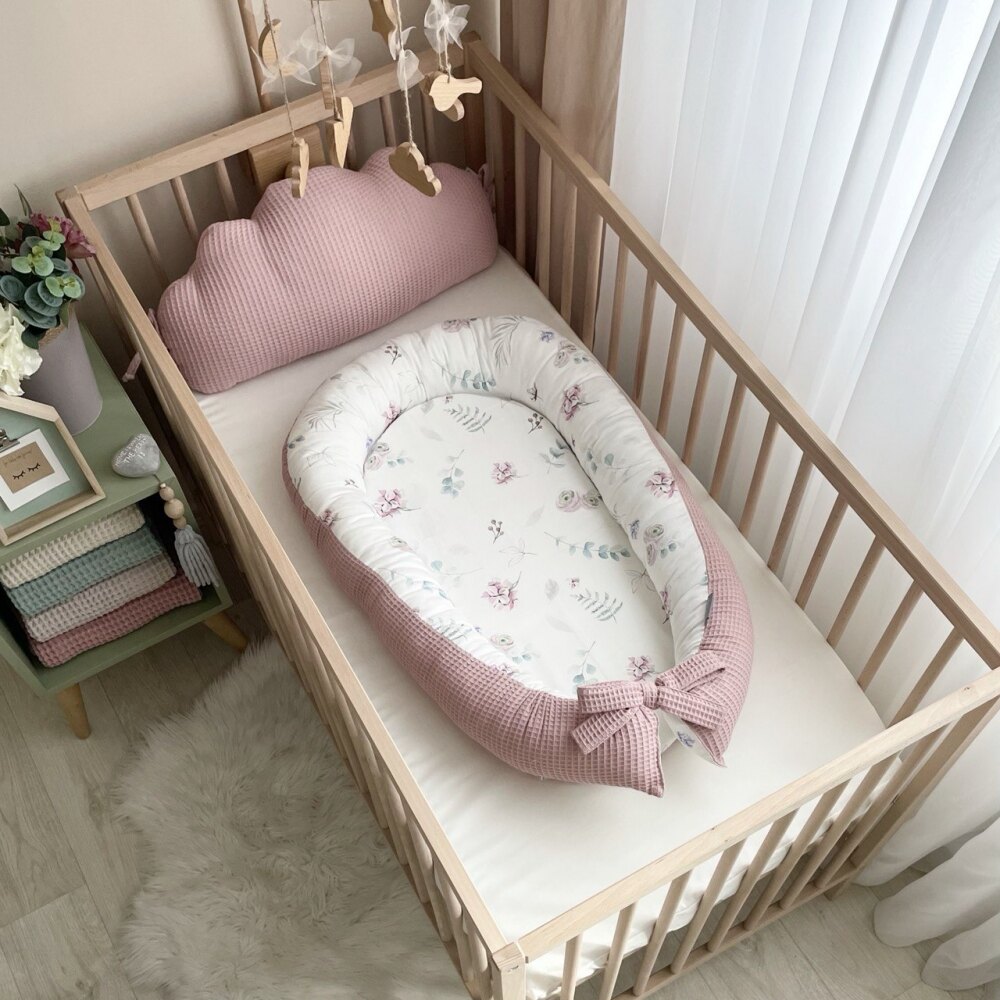 Baby nest mix de flori, roz pudră Baby nest cu flori pe fond alb și exterior din vafe roz pudră, în pătuț, lângă o pernă-nor roz