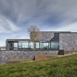 Casă modernă cu pereți din plăci Fundermax 0428 Cave, cu aspect de beton gri pătat, și un parter vitrat, pe o pantă înierbată
