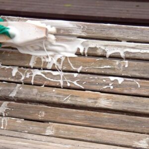 Detergent Tehopesu întins cu peria pe o terasă din lemn înnegrit de vreme, cu spumă albă prelinsă peste scânduri