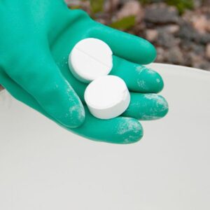 Două tablete albe Tikkurila Homeenpoisto ținute în palmă, cu mănușă verde de protecție, deasupra unei găleți albe