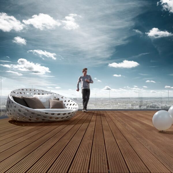 Terasă din frasin tratat termic, cu plăci maro închis riflate fin, un pat rotund împletit alb și un bărbat pe fundal de oraș